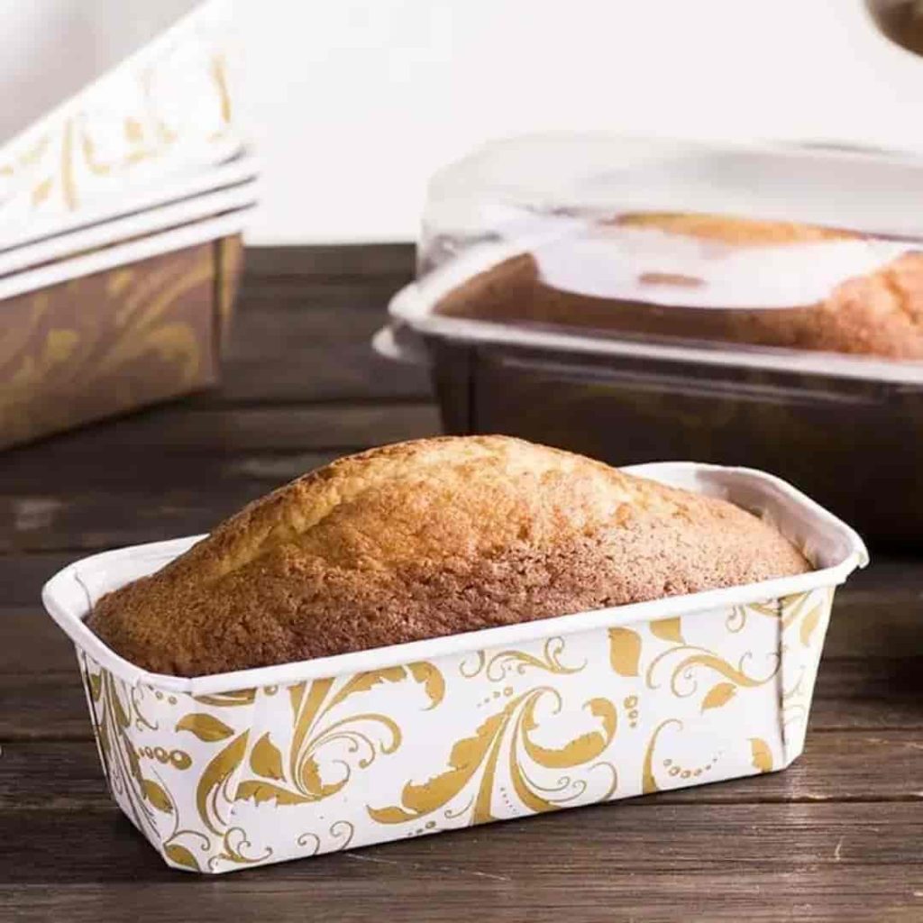 Close-up de um bolo caseiro simples assado em uma forma de papel forneável retangular com estampa decorativa em tons de dourado e branco. Ao fundo, sobre uma mesa de madeira escura, aparecem embalagens adicionais empilhadas e outro bolo protegido por tampa transparente, ilustrando a preparação para o comércio local.