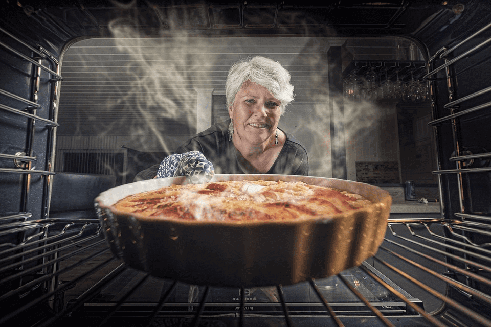 Visão de dentro do forno mostrando uma mulher loira com luva térmica retirando uma assadeira de quiche ou torta dourada, com fumaça de calor saindo do prato.