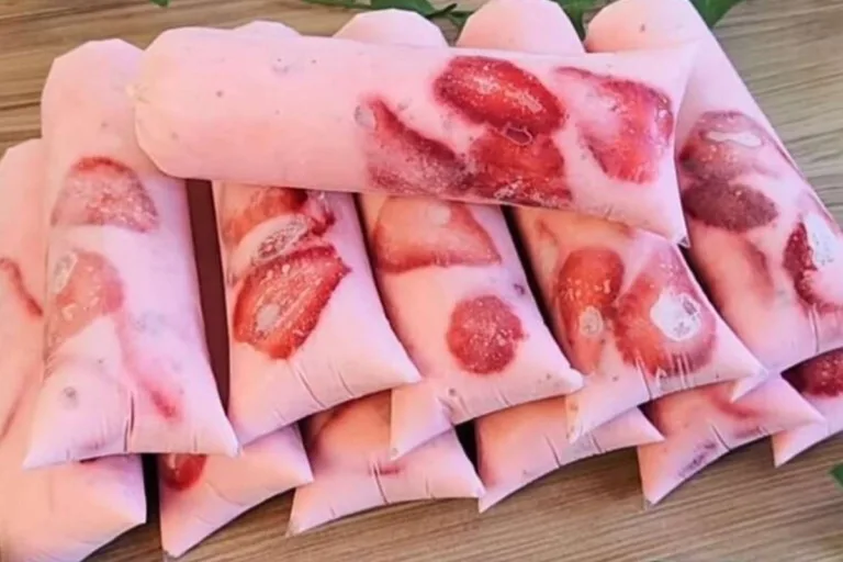 Geladinhos gourmet de morango cremoso recheados com pedaços frescos da fruta visíveis nas laterais dos saquinhos transparentes sobre uma superfície de madeira.