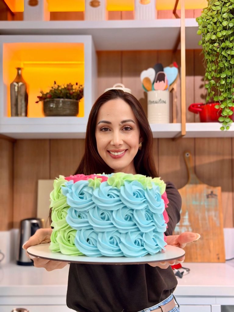 Marrara Bortoloti segurando um bolo decorado com rosetas coloridas em tons vibrantes de azul, verde e rosa.