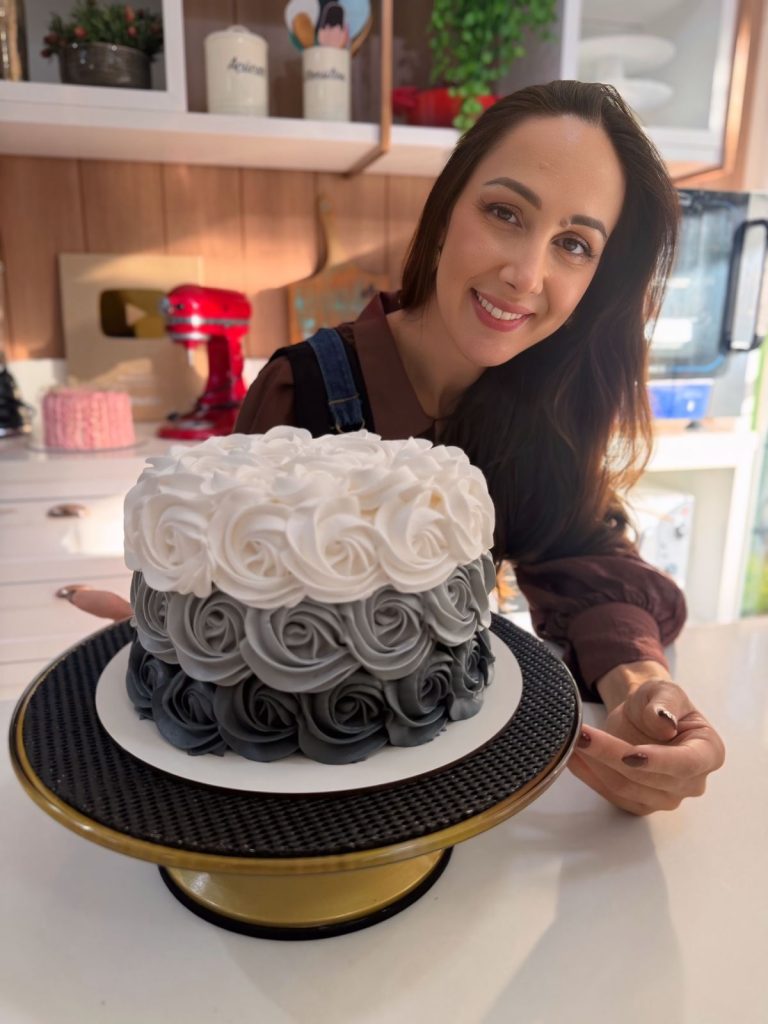 Confeiteira Marrara Bortoloti sorrindo ao lado de um bolo decorado com rosetas de chantininho em dégradé de branco, cinza e preto.