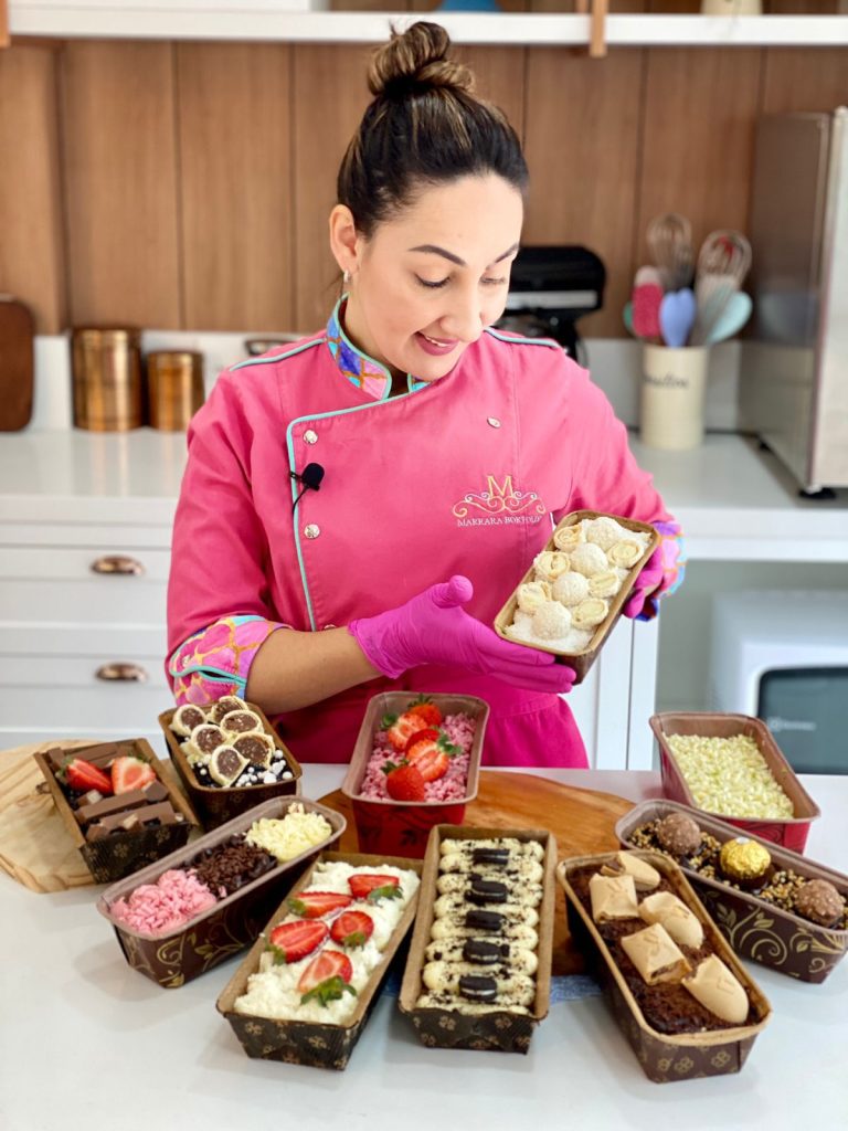 A confeiteira Marrara Bortoloti vestindo dólmã rosa com luvas, sorrindo e segurando um bolo caseirinho decorado, cercada por uma bancada repleta de diversos bolos caseirinhos em formas forneáveis.