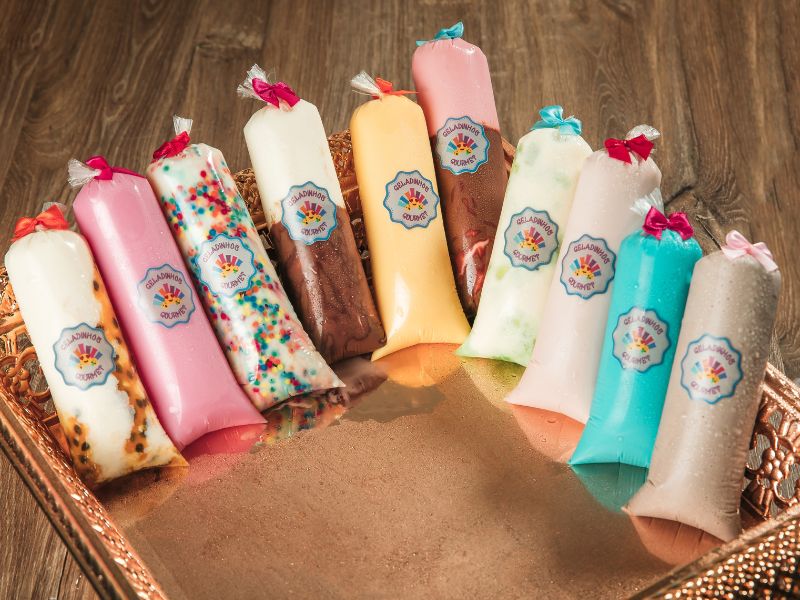 Geladinhos gourmet de diversos sabores embalados profissionalmente com etiquetas personalizadas e fitas coloridas, organizados em uma bandeja de apresentação.