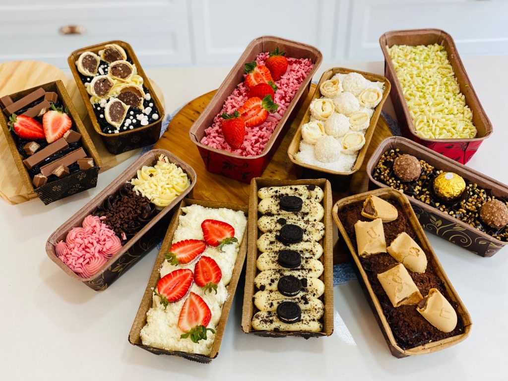 Visão superior de uma bancada exibindo uma coleção diversificada de bolos caseirinhos gourmet em formas de papel retangulares, com coberturas de morango, bombom, Oreo, coco e chocolate.