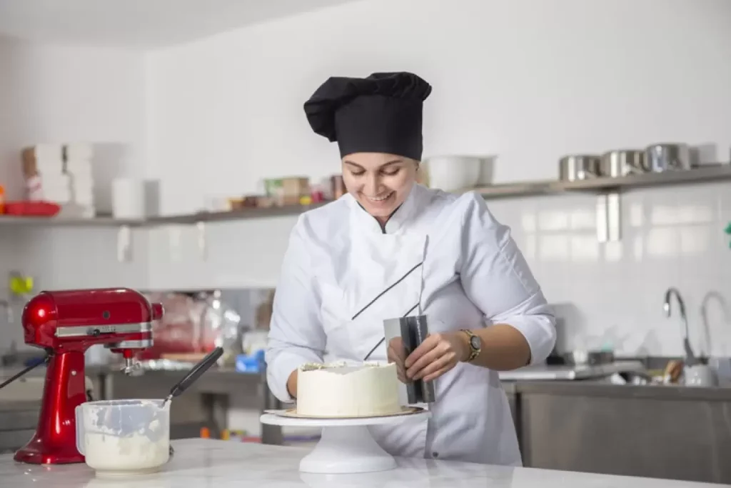 Confeiteira sorridente com uniforme branco e toque blanche, utilizando uma espátula para nivelar a cobertura de um bolo branco em um suporte giratório em uma cozinha profissional equipada.