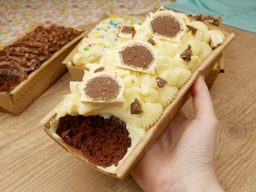 Close-up de uma mão segurando um "bolo caseirinho" gourmet de chocolate com cobertura branca e trufas cortadas no topo, servido em forma de papel forneável. Ao fundo, desfocadas, outras unidades demonstram produção em escala. A imagem ilustra o produto final de alto valor que precisa ter seus custos calculados com precisão.