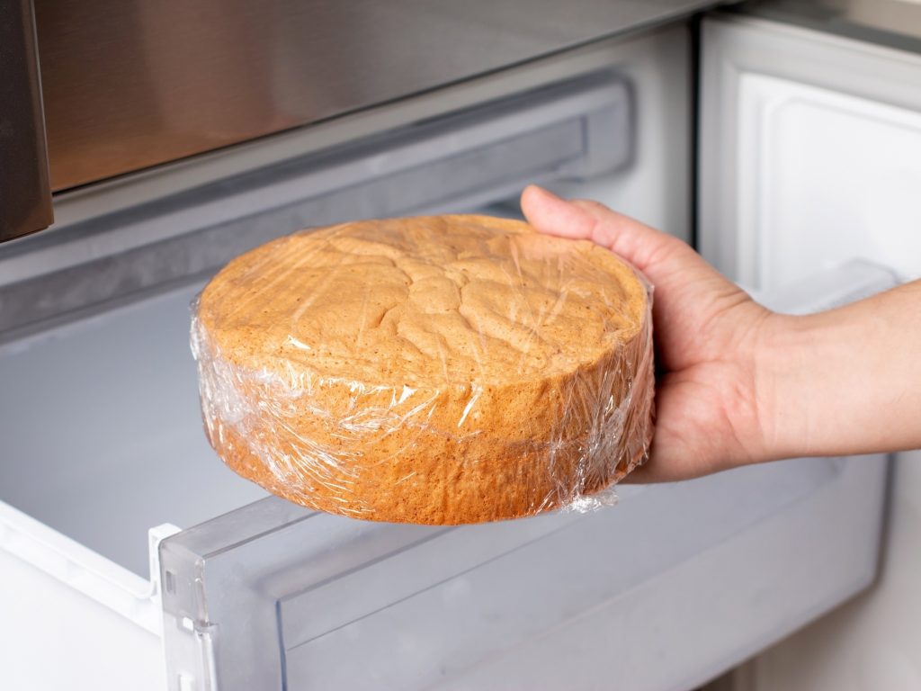 Close-up de uma mão segurando uma massa redonda de bolo simples, firmemente embalada em filme plástico transparente, prestes a ser inserida na gaveta aberta de um freezer doméstico.