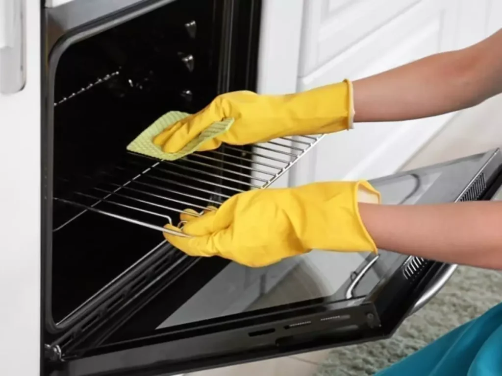 Close de mãos usando luvas amarelas de proteção limpando as grades internas de um forno elétrico branco com um pano verde.