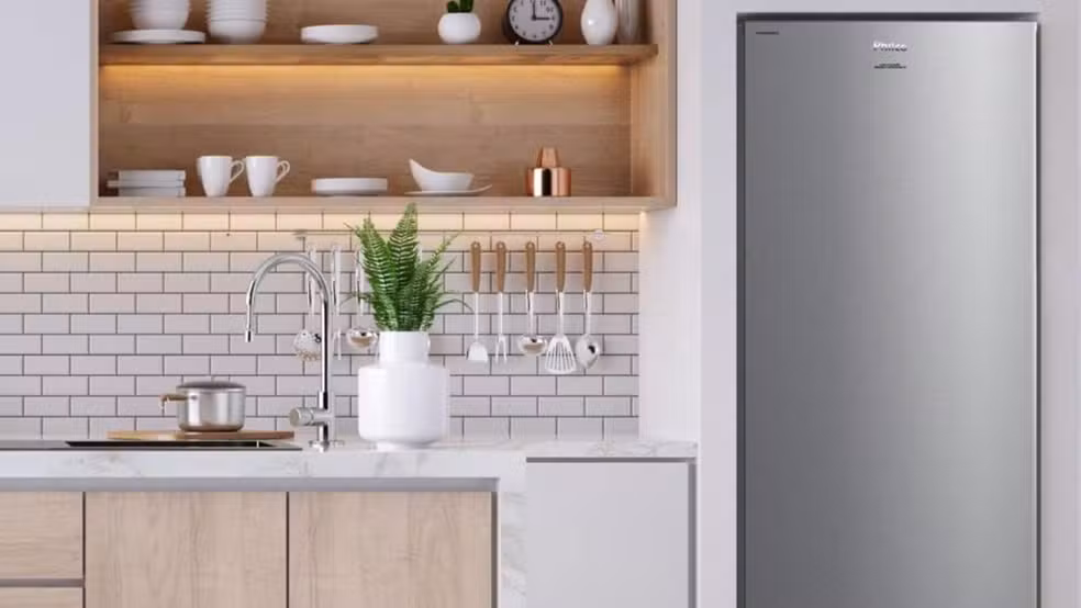 Freezer vertical Philco em acabamento inox posicionado em uma cozinha moderna com revestimento de tijolos brancos, bancada de mármore e prateleiras de madeira iluminadas.