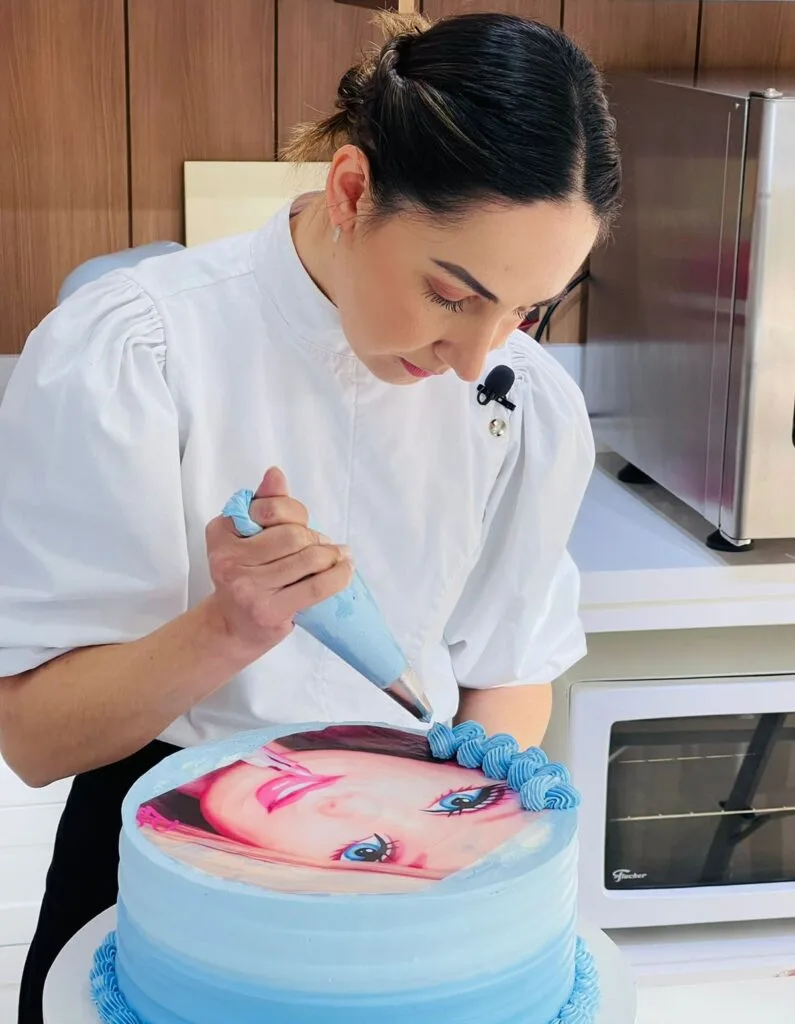Marrara Bortoloti aplicando bico de confeitar em um bolo de aniversário rosa com tema de borboletas, mostrando uma das técnicas ensinadas no curso.