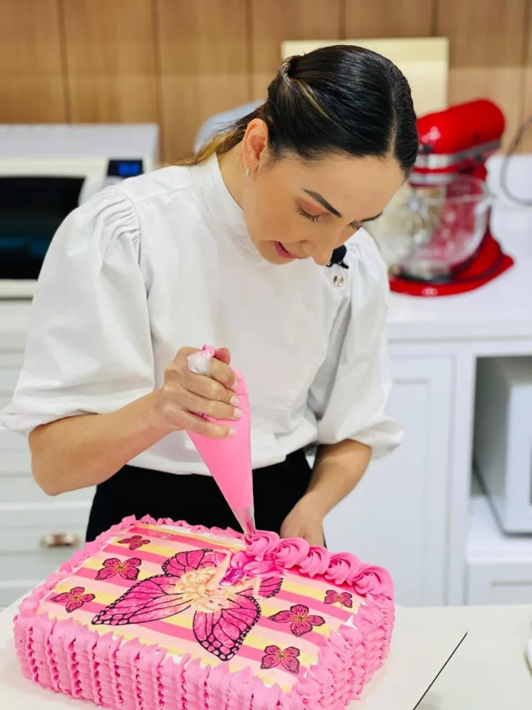 Marrara Bortoloti decorando um bolo de princesa com bico de confeitar e cobertura azul, demonstrando uma das técnicas de finalização.