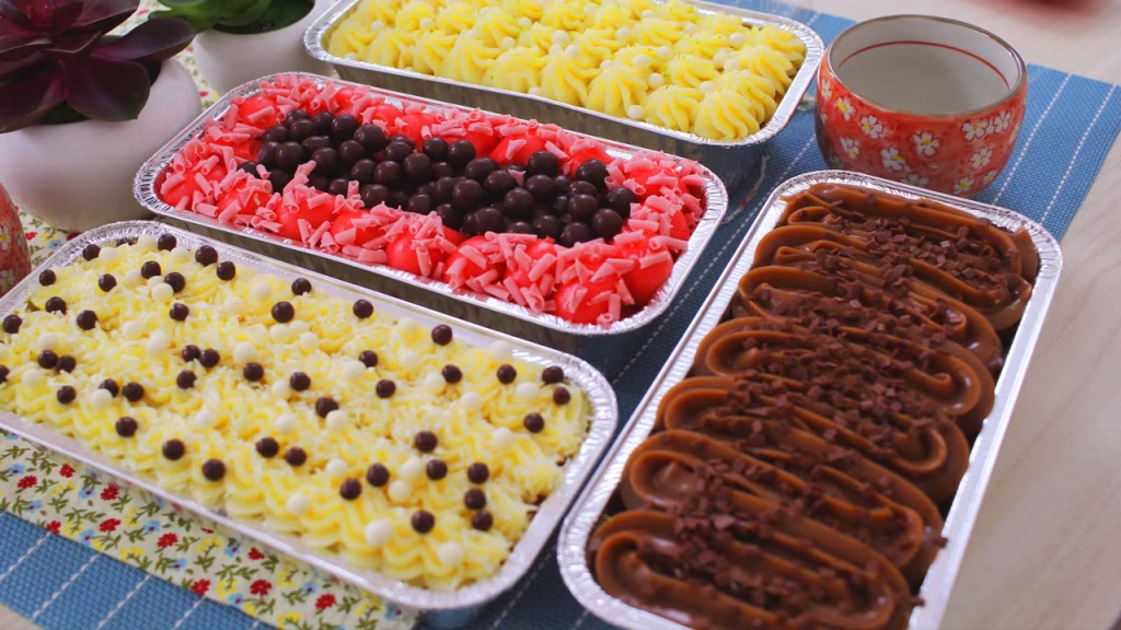 Visão de close-up de quatro variedades de "bolos caseirinhos" gourmet dispostos ordenadamente em formas de alumínio descartáveis sobre uma toalha de mesa com estampa floral azul. A imagem exibe produtos finalizados e estruturados: duas versões com cobertura amarela (uma com granulado, outra com pérolas brancas), uma com cobertura vermelha e bolinhas de chocolate, e uma com cobertura marrom de brigadeiro. Eles demonstram a estabilidade da massa e a ausência de deformações nas bordas da embalagem.