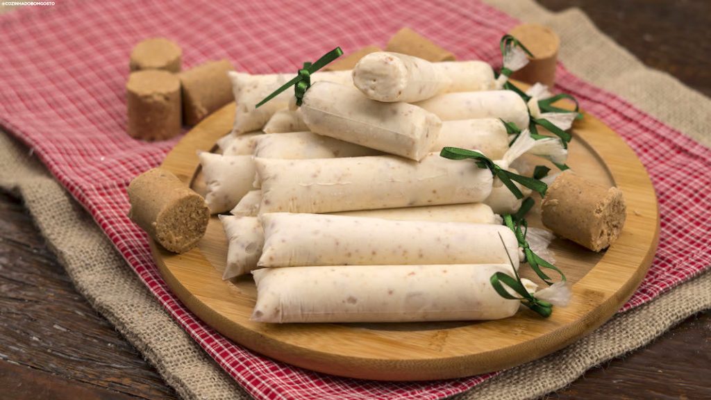 Geladinhos gourmet de paçoca artesanais amarrados com fitas verdes e decorados com paçocas rolha inteiras sobre uma tábua de madeira e tecido xadrez.