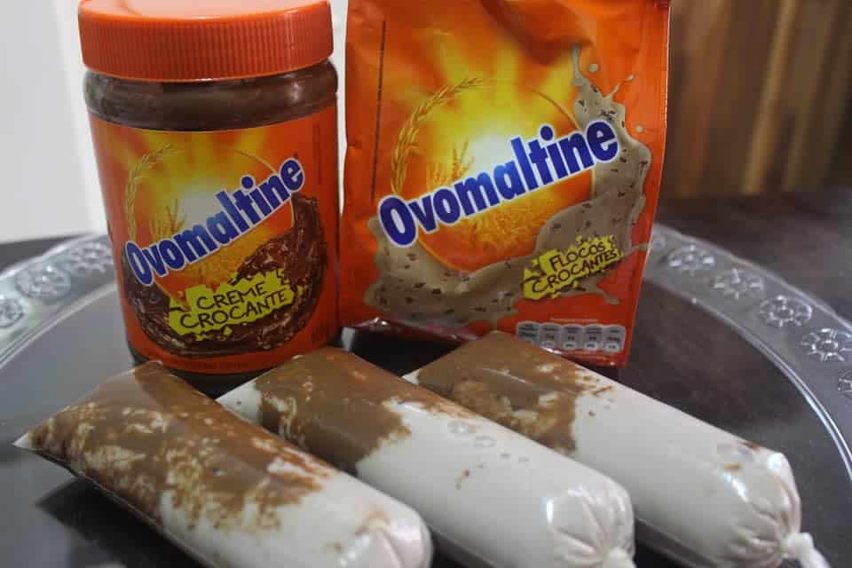 Geladinhos gourmet de Ovomaltine artesanais dispostos ao lado do pote de creme crocante e da embalagem oficial do ingrediente em uma bandeja.