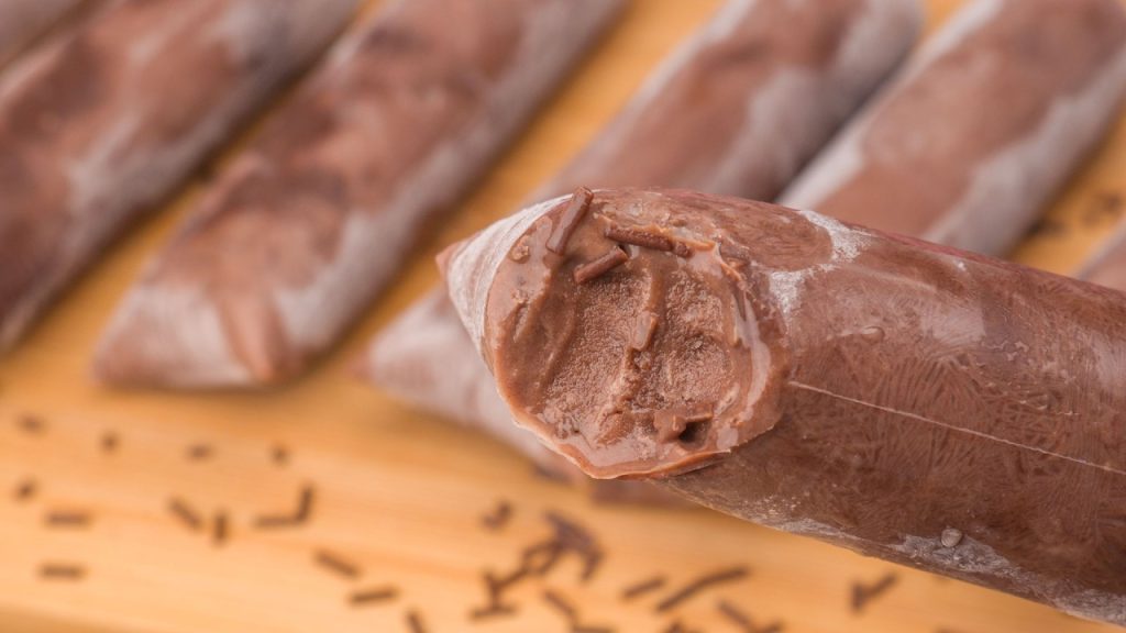 Geladinho gourmet de brigadeiro aberto mostrando textura cremosa e granulado de chocolate, com outros geladinhos artesanais ao fundo sobre uma tábua de madeira.