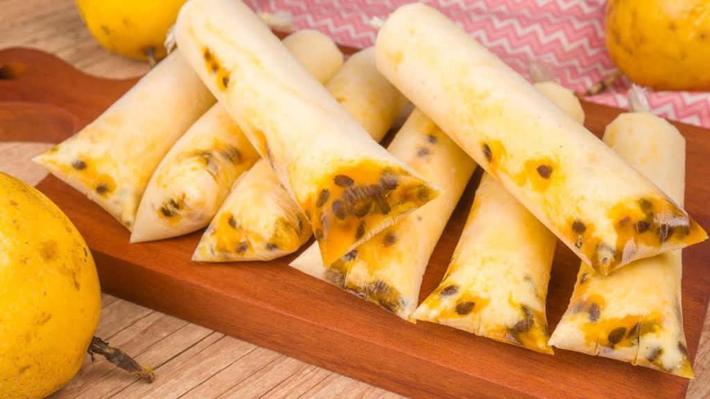 Geladinhos gourmet de maracujá cremoso com calda artesanal e sementes visíveis nas extremidades, dispostos sobre uma tábua de madeira ao lado de frutos inteiros.