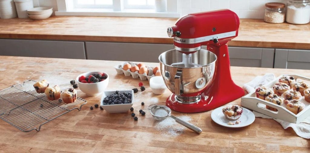 Batedeira planetária vermelha KitchenAid em bancada de madeira, cercada por ingredientes e muffins de frutas. Acessórios e tigela de inox em destaque.