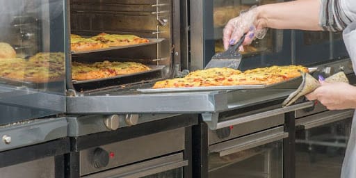 Close das mãos de uma profissional retirando uma assadeira de pizzas douradas de um forno industrial com múltiplas câmaras e calor estático.