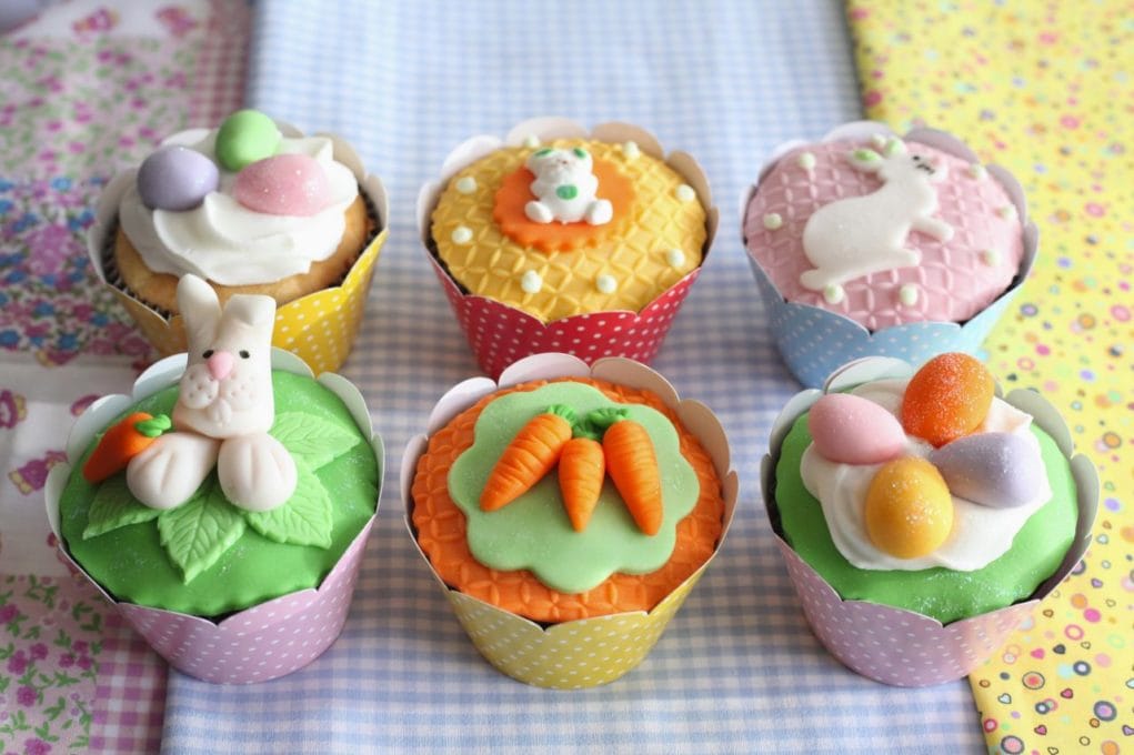 Seis cupcakes de Páscoa em forminhas coloridas, decorados com coelhos de fondant, ovos coloridos e cenouras sobre uma toalha de mesa quadriculada.