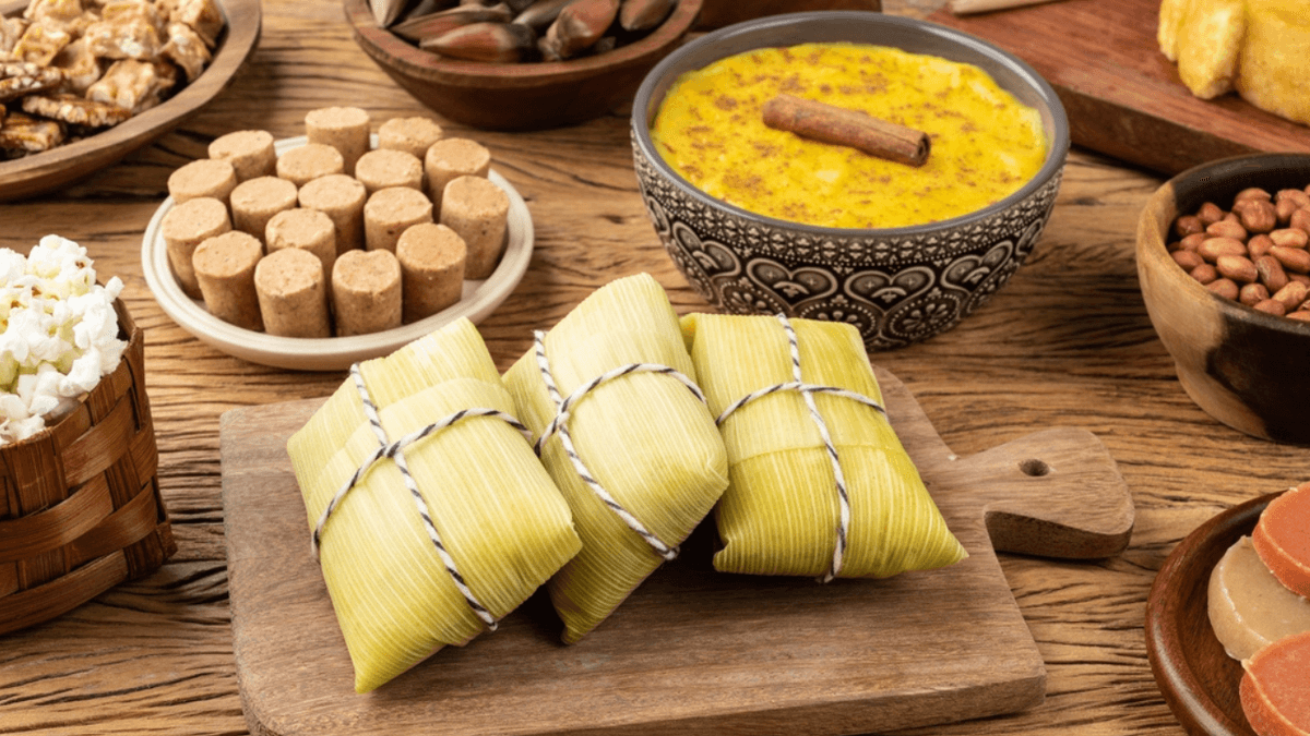 Mesa de madeira com três pamonhas amarradas em palha de milho, um pote de curau com canela, paçocas, amendoins e pipoca, representando doces de Festa Junina.
