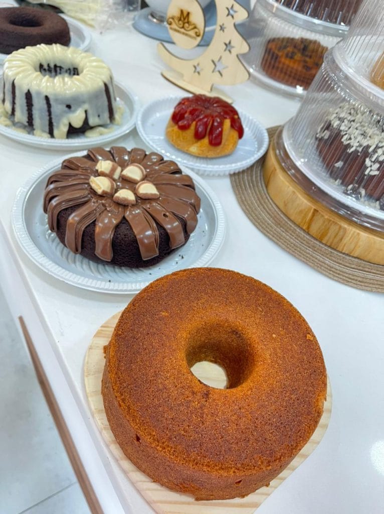 Uma mesa cheia de bolos caseiros de diferentes sabores e coberturas, incluindo chocolate com granulado, coco ralado, doce de leite e chocolate branco.