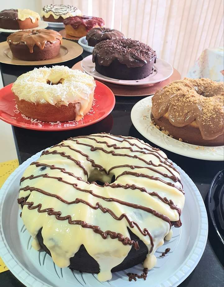 Uma mesa cheia de bolos caseiros com diferentes coberturas, incluindo chocolate, coco e calda de doce de leite