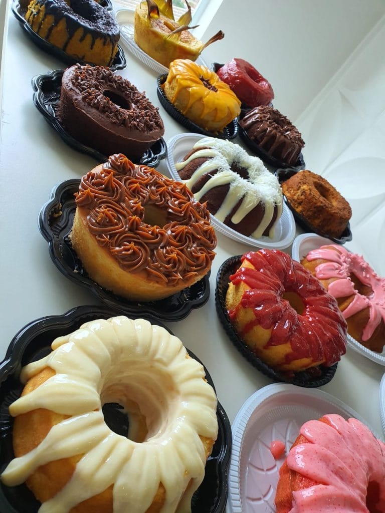 ma mesa com diversos bolos caseiros com diferentes coberturas, incluindo chocolate, coco, caramelo, frutas vermelhas e rosa, dispostos em pratos.