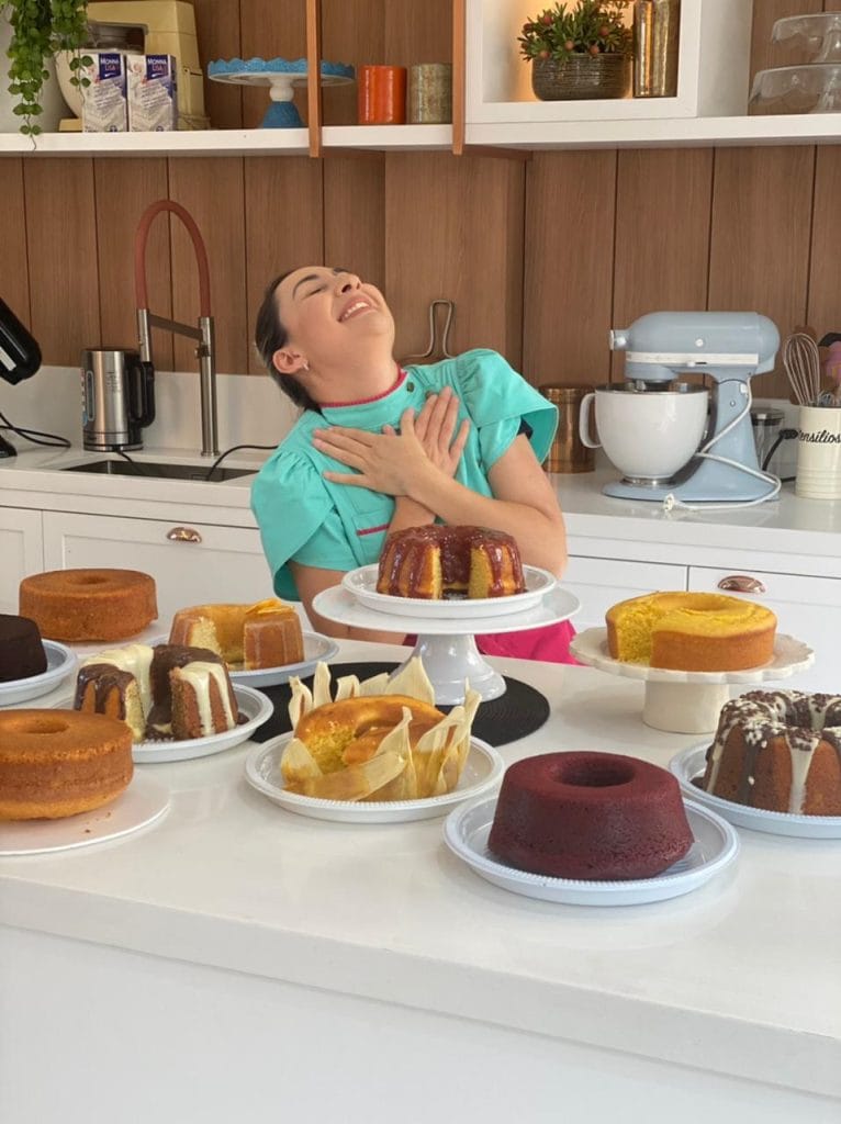 A confeiteira Marra Bortolotti em uma cozinha, sorrindo e com a mão no peito, cercada por uma mesa cheia de diversos bolos caseiros.