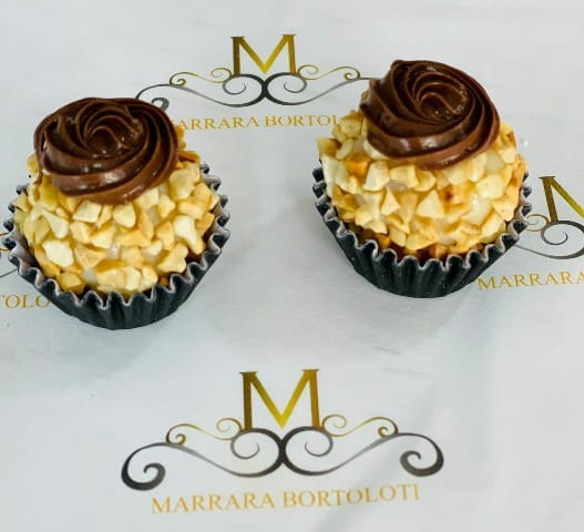 Dois brigadeiros gourmet em forminhas pretas, cobertos com pedaços de amendoim e um toque de ganache de chocolate no topo, sobre um fundo branco com a marca "Marrara Bortoloti".
