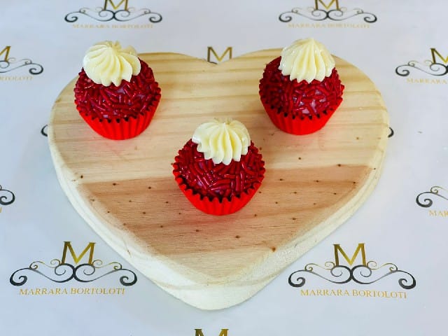 Três docinhos de festa em forminhas vermelhas, cobertos com granulado vermelho e um pequeno tufo de creme branco no topo, dispostos sobre um suporte de madeira em formato de coração.