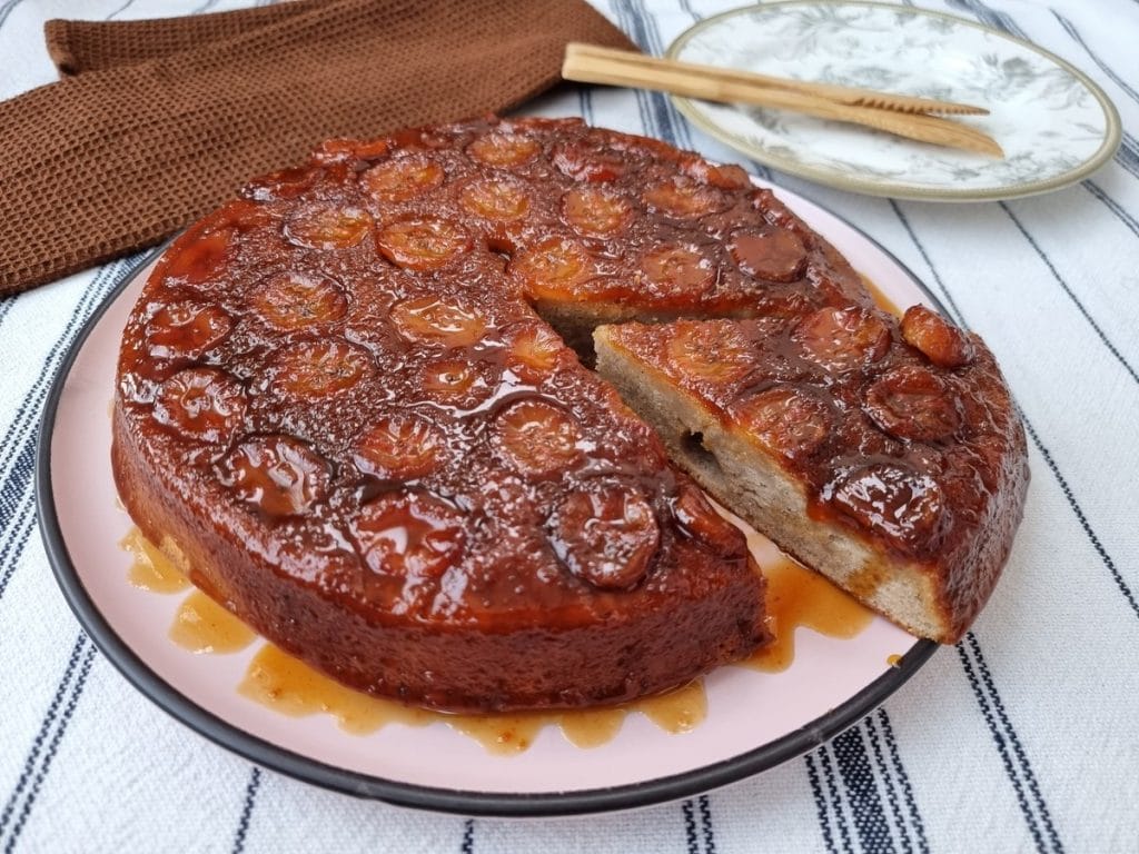 Um bolo de banana redondo com uma camada brilhante de caramelo e fatias de banana no topo, com uma fatia cortada mostrando a massa clara e fofinha.