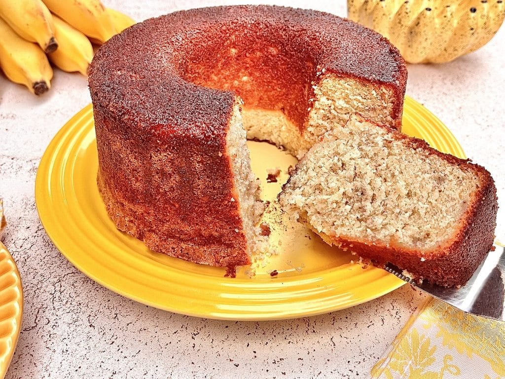 Um bolo de banana fofinho com formato de anel, em um prato amarelo, com uma fatia cortada ao lado que revela a textura macia do interior.