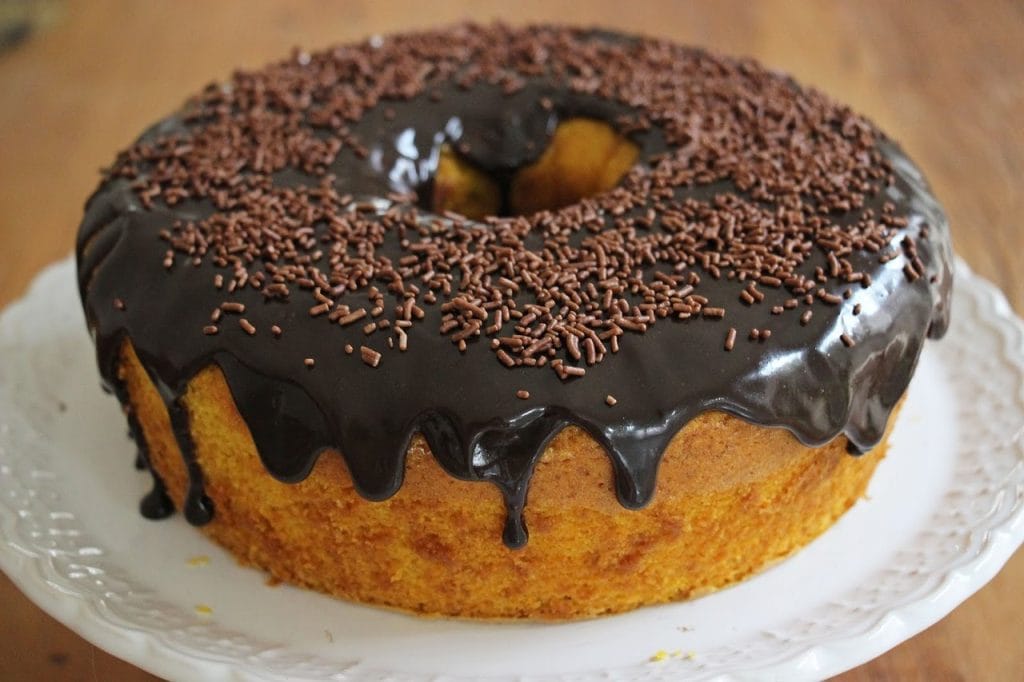 Um bolo de cenoura caseiro em formato de anel, com uma generosa e brilhante cobertura de chocolate e granulados de chocolate por cima.
