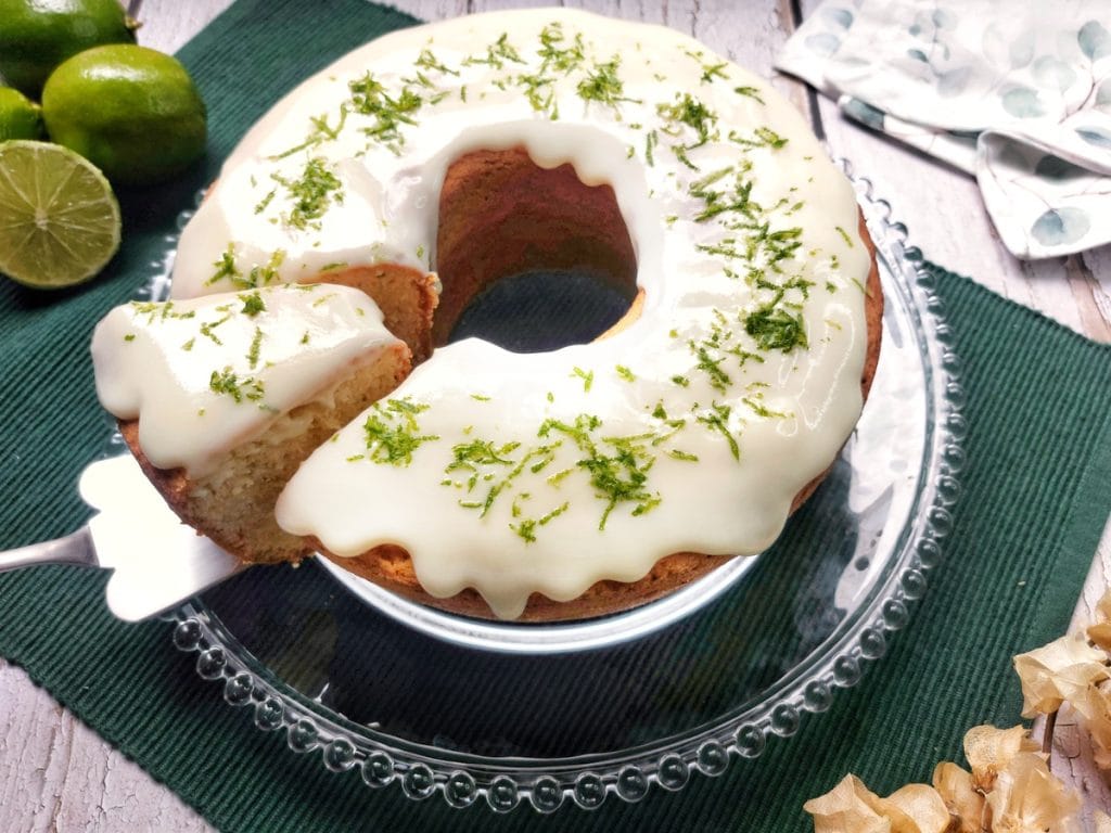 Um bolo de limão em forma de anel com uma cobertura branca cremosa e raspas de limão por cima, sobre um suporte de vidro, com uma fatia já cortada.