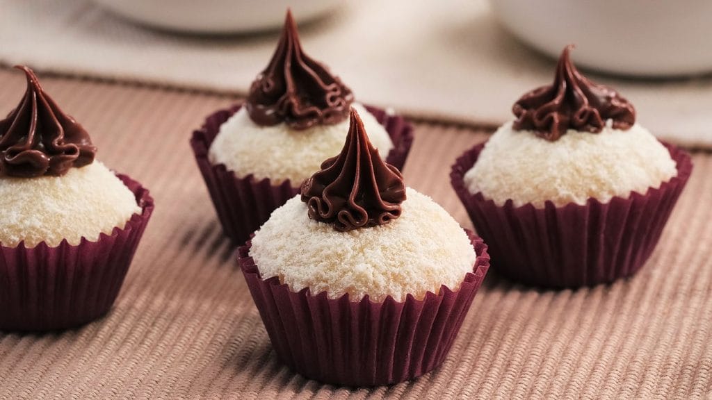 Quatro brigadeiros de Leite Ninho, cobertos com um recheio de Nutella, em forminhas de papel na cor bordô, dispostos sobre uma toalha rosa.
