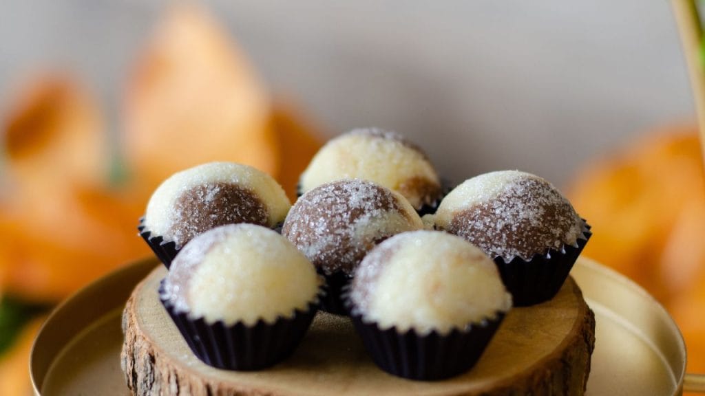 Seis docinhos casadinhos de chocolate e beijinho, cobertos com açúcar cristal, em um suporte de madeira sobre uma bandeja dourada.