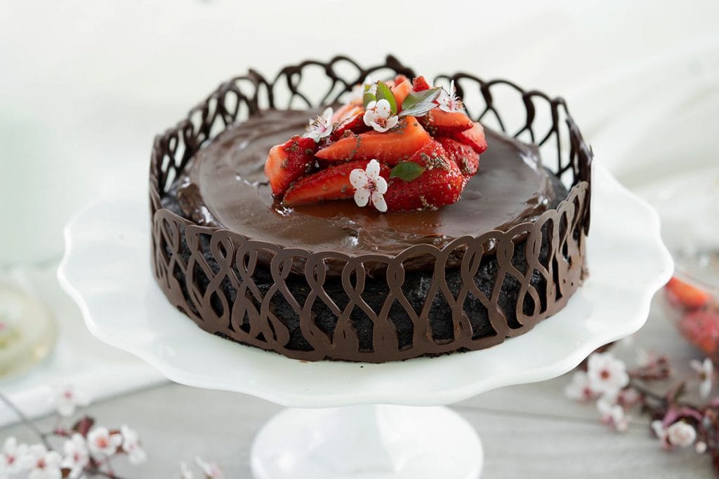 Bolo de chocolate coberto com ganache brilhante, cercado por uma barreira de chocolate e decorado com morangos frescos e flores no topo.