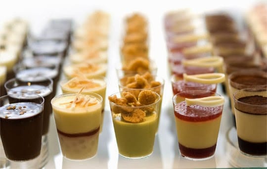 Uma bandeja com várias fileiras de doces em pequenos copinhos transparentes, com diferentes recheios e coberturas, como mousse, calda de frutas e granulados.