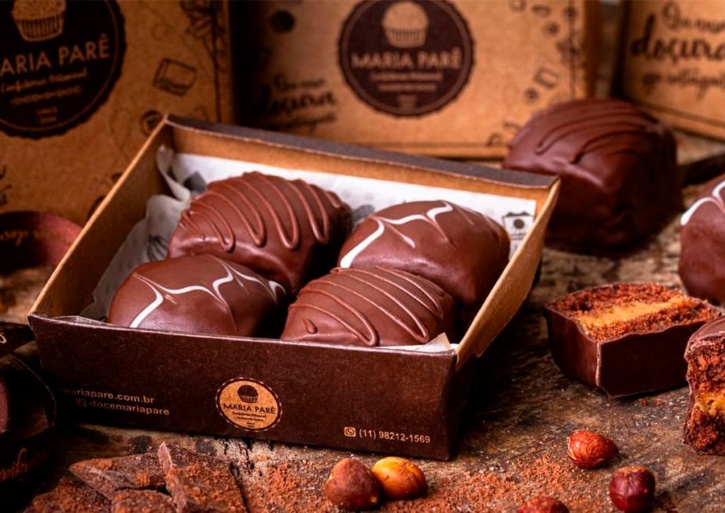 Uma caixa marrom de papelão com quatro doces de chocolate com decorações em chocolate branco, sobre um fundo rústico com lascas de chocolate, avelãs e outras caixas da mesma marca.
