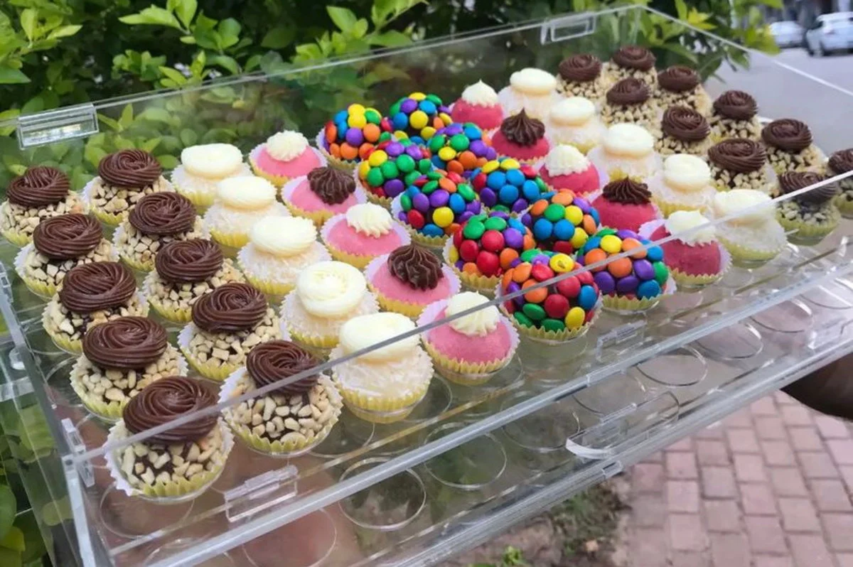 Uma bandeja de acrílico transparente contendo diversos tipos de docinhos de festa coloridos, prontos para venda na rua.