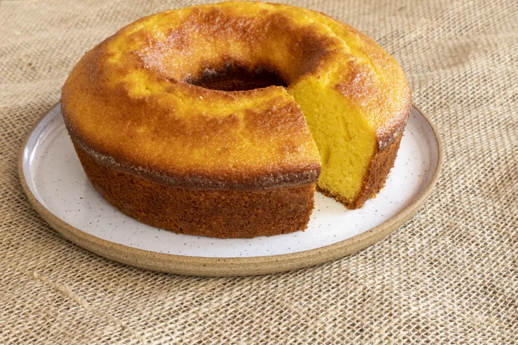 Um bolo de laranja com uma fatia cortada, mostrando a massa amarela e fofinha. O bolo está em um prato de cerâmica rústico sobre uma superfície de estopa.