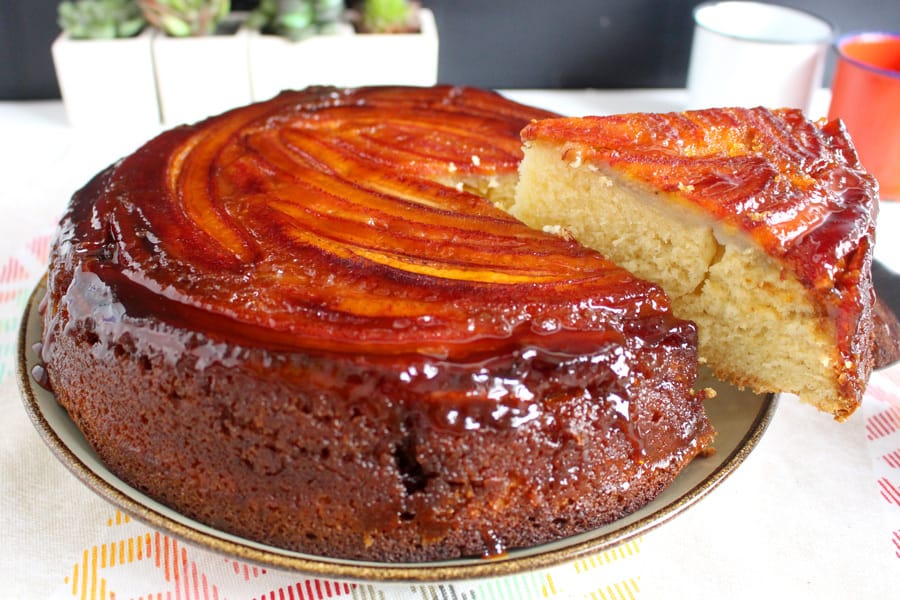 Um bolo de banana redondo com uma cobertura brilhante de caramelo e fatias de banana dispostas em espiral no topo, com uma fatia cortada e ao lado, mostrando o miolo fofinho.