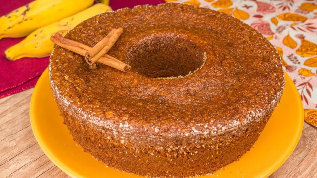Um bolo de banana fofinho em um prato amarelo, com um buraco no meio e decorado com dois paus de canela no topo.