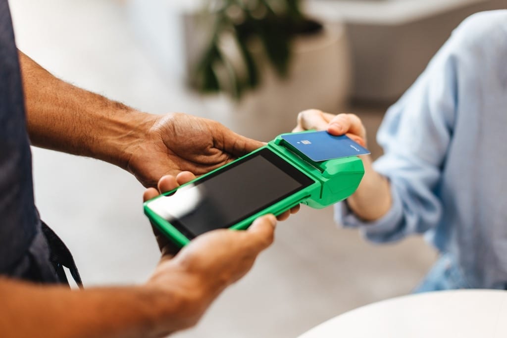Mãos segurando uma maquininha de cartão verde, enquanto um cliente aproxima um cartão de crédito azul para realizar um pagamento por aproximação.