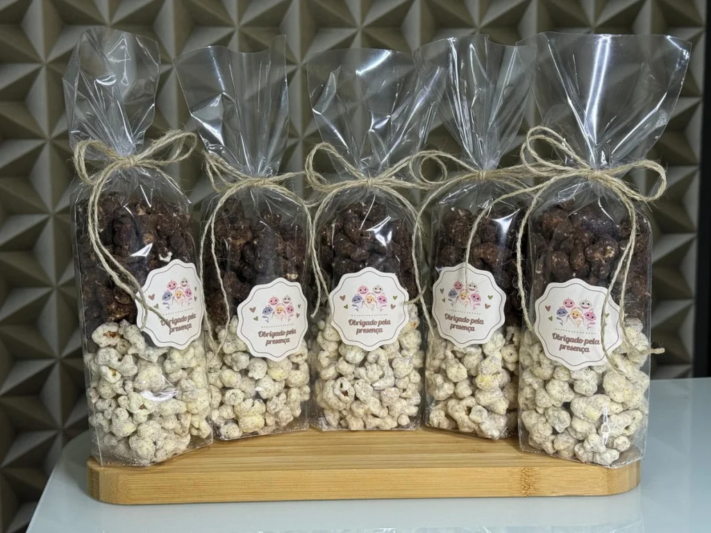 Cinco pacotes transparentes de pipoca gourmet, com camadas de pipoca caramelizada escura e pipoca doce branca, amarrados com barbante e com uma etiqueta de agradecimento, sobre uma tábua de madeira.