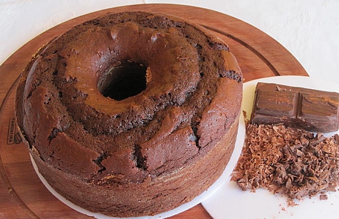 Um bolo de chocolate simples e caseiro em formato de anel, com uma barra de chocolate e raspas de chocolate ao lado, sobre um prato branco.