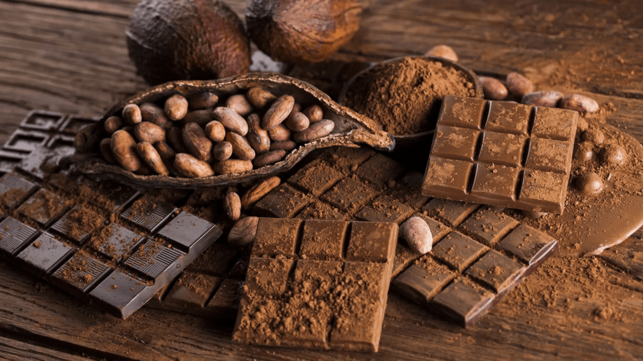 Imagem de destaque: variedade de chocolates em pó, barras de chocolate ao leite e amargo, cacau em pó e grãos de cacau em uma superfície de madeira rústica, destacando a riqueza dos ingredientes para confeitaria.