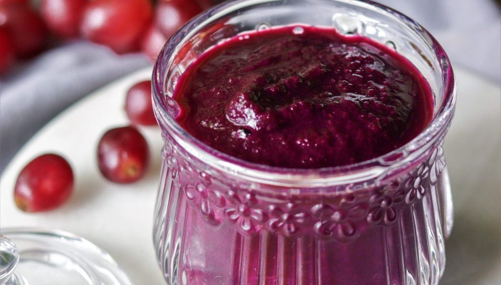 Pote de vidro com geleia de uva caseira escura e brilhante, decorado com um tecido xadrez e fita, pronto para ser armazenado ou servido. Destaca a embalagem e a conservação.