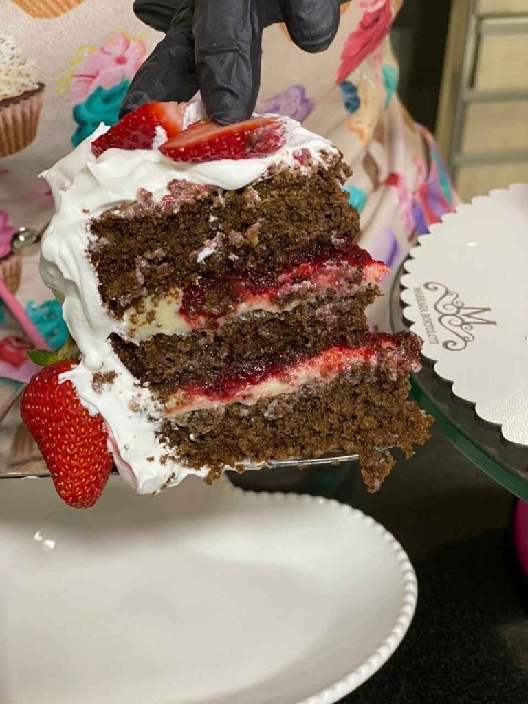Uma fatia de bolo de chocolate com camadas de recheio cremoso, geleia de morango e chantilly, sobre uma espátula, com morangos frescos ao lado.