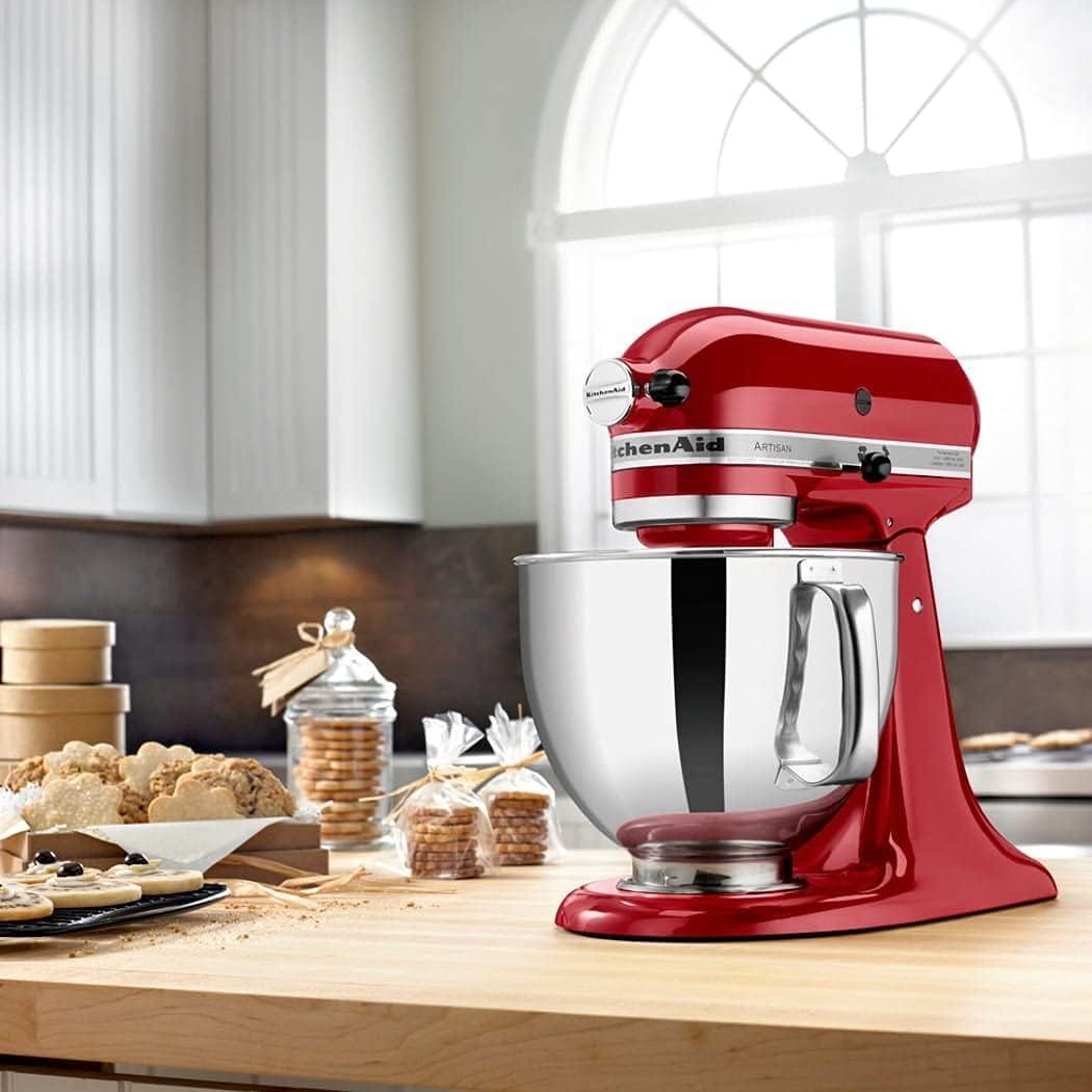 Batedeira KitchenAid Stand Mixer Artisan Vermelha (Empire Red) 220V em uma bancada de madeira, cercada por biscoitos recém-assados e outros doces, ilustrando seu uso em uma confeitaria.